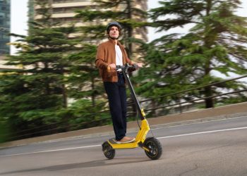 Xiaomi Electric Scooter 6 Ultra em cor amarela estacionada numa rua urbana, ao lado do Xiaomi Watch 5 no pulso de um utilizador