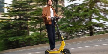 Xiaomi Electric Scooter 6 Ultra em cor amarela estacionada numa rua urbana, ao lado do Xiaomi Watch 5 no pulso de um utilizador