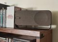 Análise Bose SoundLink Home: quando o design soa mais alto 11 Bose SoundLink Home em Light Silver sobre prateleira de madeira clara, com a grelha de tecido e a banda metálica contínua em destaque num ambiente de sala minimalista