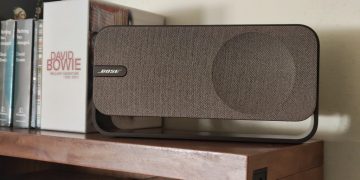 Bose SoundLink Home em Light Silver sobre prateleira de madeira clara, com a grelha de tecido e a banda metálica contínua em destaque num ambiente de sala minimalista