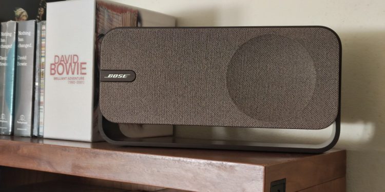 Análise Bose SoundLink Home: quando o design soa mais alto 1 Bose SoundLink Home em Light Silver sobre prateleira de madeira clara, com a grelha de tecido e a banda metálica contínua em destaque num ambiente de sala minimalista