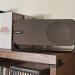 Análise Bose SoundLink Home: quando o design soa mais alto 6 Bose SoundLink Home em Light Silver sobre prateleira de madeira clara, com a grelha de tecido e a banda metálica contínua em destaque num ambiente de sala minimalista